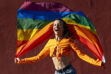 Happy young woman in casual clothes with rainbow flag up against terracotta wall on city street - ADSF39869