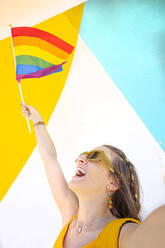 Glückliche Lesbe in Freizeitkleidung, die den Arm mit der Regenbogenfahne hebt und nach oben schaut, während sie den Monat des Stolzes an einer bunten Wand auf der Straße feiert - ADSF39871
