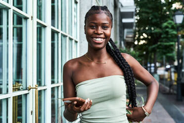 Happy African American woman with long braids wearing stylish tube top and browsing smartphone on city street - ADSF39899