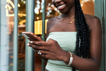 Crop African American female in stylish top smiling and sending text message on smartphone while leaning on glass wall on city street - ADSF39901