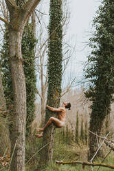 Side view full body of tranquil male with nude body and closed eyes hugging tree trunk in woods in summer - ADSF39913