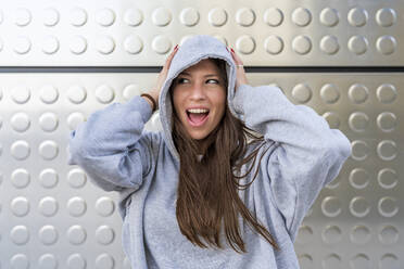 Woman with mouth open wearing hooded shirt in front of metal wall - DLTSF03208