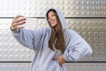Smiling woman taking selfie through smart phone in front of metal wall - DLTSF03211