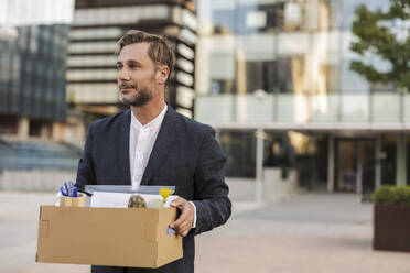 Businessman carrying cardboard box outside office building - JCCMF07706