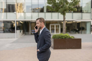 Geschäftsmann, der mit seinem Smartphone vor einem Bürogebäude spricht - JCCMF07707