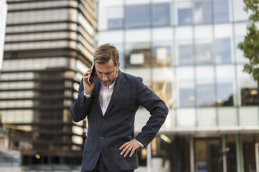 Tensed businessman talking on smart phone in front of office building - JCCMF07711