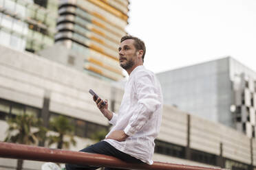 Nachdenklicher Mann sitzt mit Smartphone auf dem Geländer - JCCMF07714