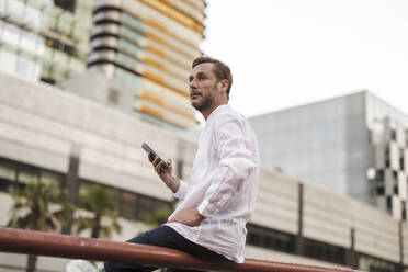 Thoughtful man sitting with smart phone on railing - JCCMF07714