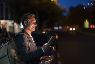 Smiling businessman with backpack holding smart phone at street - JCCMF07739