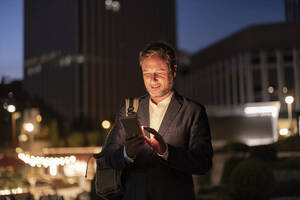 Smiling businessman using mobile phone in city at night - JCCMF07740