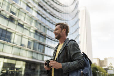 Smiling man with camera standing in front of building - JCCMF07748