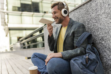 Smiling man talking on speaker phone sitting by wall - JCCMF07753