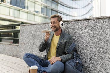 Happy man talking on speaker phone sitting by wall - JCCMF07754