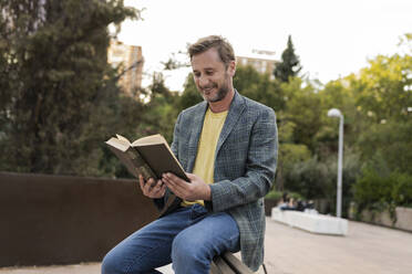 Happy man reading book sitting on bench at park - JCCMF07755