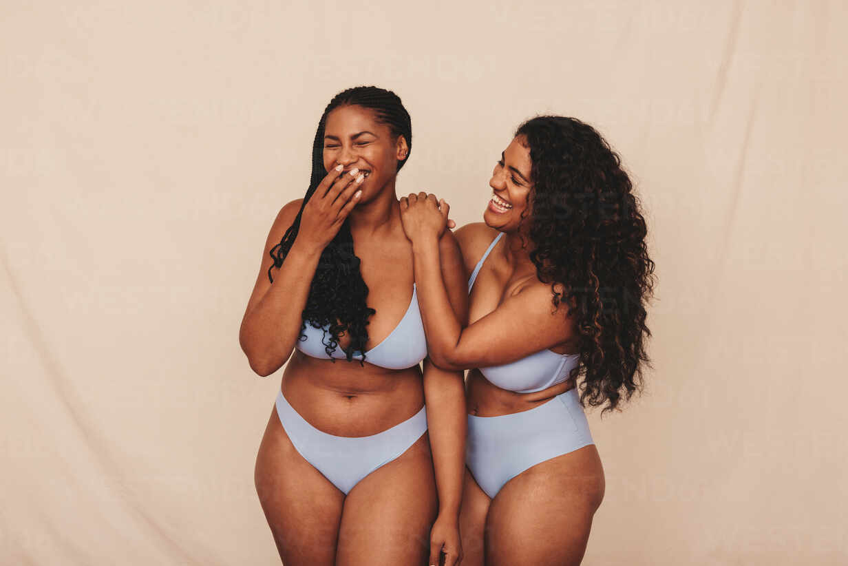 Cheerful young women laughing in blue underwear. Two happy young women  celebrating their natural bodies and curves in a studio. Body positive  young women standing together against a studio background. stock photo