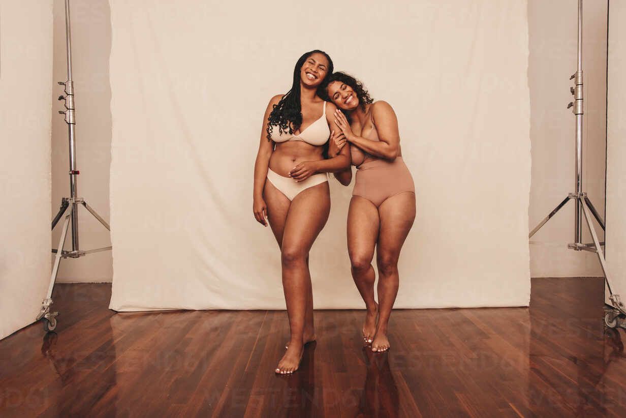 Young women embracing their natural bodies in a studio. Two happy young  women smiling cheerfully while standing in underwear against a studio  background. Women feeling comfortable in their bodies. stock photo