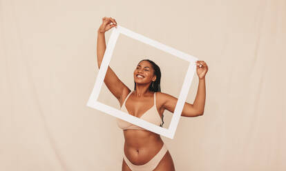 Playful young woman smiling through a picture frame in a studio. Happy young woman wearing beige underwear against a studio background. Young woman embracing her natural body and beauty. - JLPSF12132