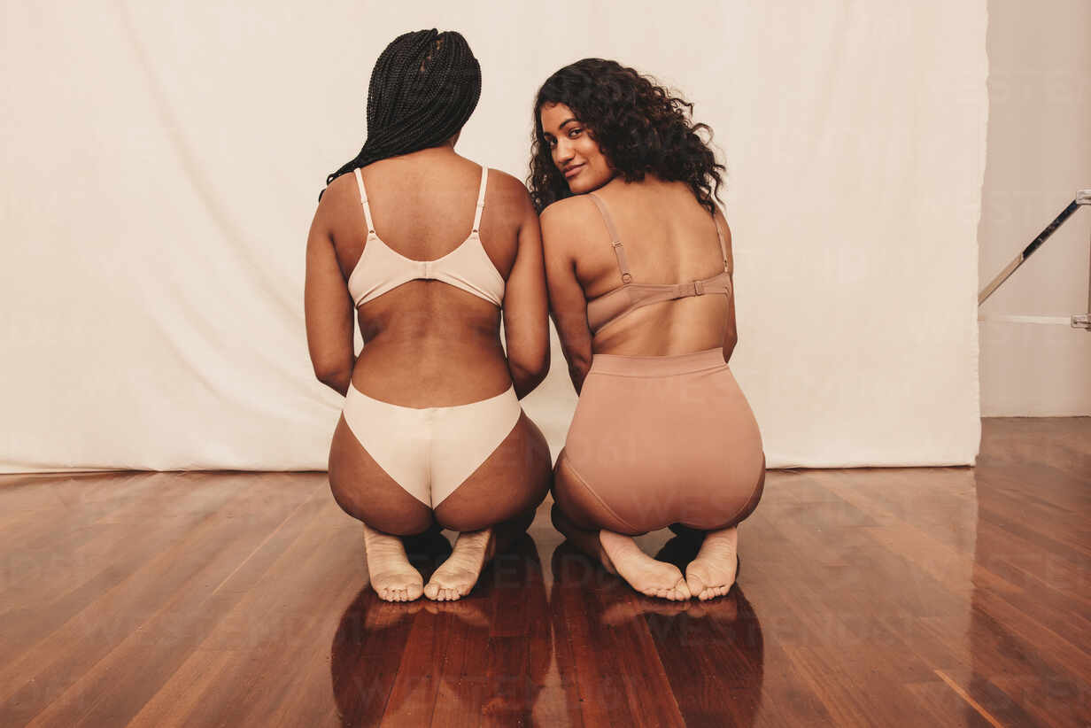Two confident young women kneeling in underwear. Two body positive young  women feeling comfortable in their natural bodies. Female friends embracing  their own bodies in a studio. stock photo