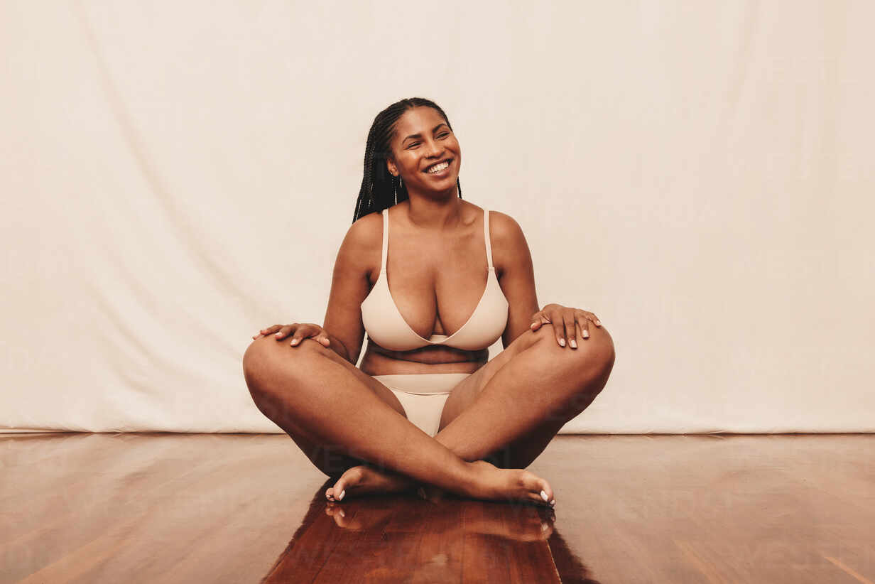 Body positivity and happiness. Young woman smiling cheerfully while sitting  with her legs crossed in a studio. Confident young woman embracing her  natural body while wearing beige underwear. stock photo