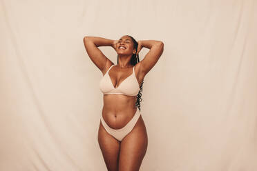 Soaking in self love. Happy young woman smiling cheerfully while standing against a studio background. Confident young woman embracing her natural body and beauty while wearing beige underwear. - JLPSF12148