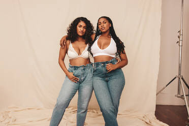 Zwei selbstbewusste junge Frauen schauen in die Kamera, während sie zusammen vor einem Studiohintergrund stehen. Zwei Freundinnen, die ihre natürlichen Körper umarmen. - JLPSF12191