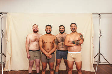 Happy and shirtless. Four body positive men laughing cheerfully while  standing together in underwear. Self-confident young men embracing their  natural bodies in a studio. stock photo