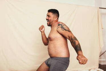 Celebrating being natural. Happy shirtless man dancing cheerfully while wearing grey underwear and standing against a studio background. Body positive and self-confident man embracing his own body. - JLPSF12251