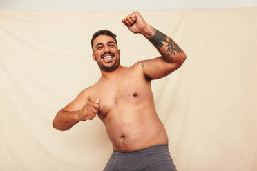 Man dancing cheerfully in the studio. Happy shirtless man celebrating while standing against a studio background. Body positive and self-confident man embracing his natural body. - JLPSF12253