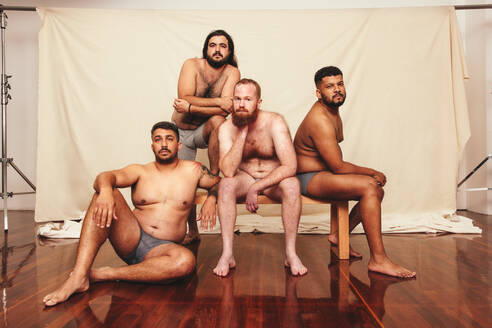 Ohne Hemd und selbstbewusst. Vier körperbewusste Männer, die in Unterwäsche in einem Studio in die Kamera schauen. Eine Gruppe selbstbewusster Männer, die ihre natürlichen Körper gemeinsam umarmen. - JLPSF12257