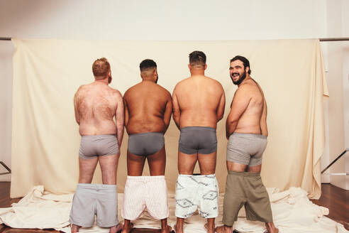 Four body positive men standing in a studio with their shorts dropped down. Rearview of a group of men standing together in underwear. Self-confident men flexing their natural bodies. - JLPSF12261