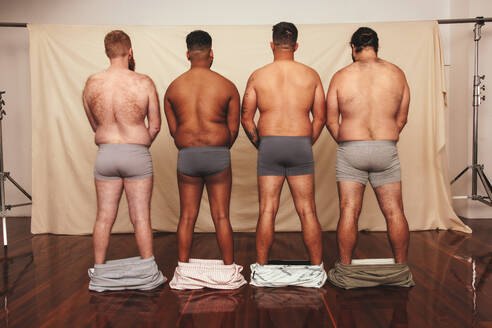 Unrecognizable men standing with their shorts dropped down. Rearview of a group of body positive men standing together in underwear. Self-confident men flexing their natural bodies. - JLPSF12263