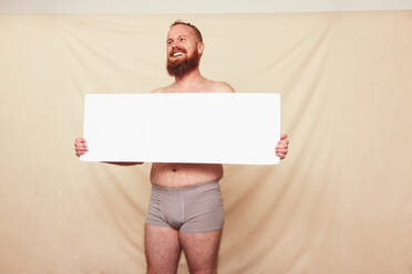 Advocating for body positivity and natural bodies. Happy shirtless man smiling cheerfully while displaying a blank white placard in a studio. Self-confident man standing in grey underwear. - JLPSF12270