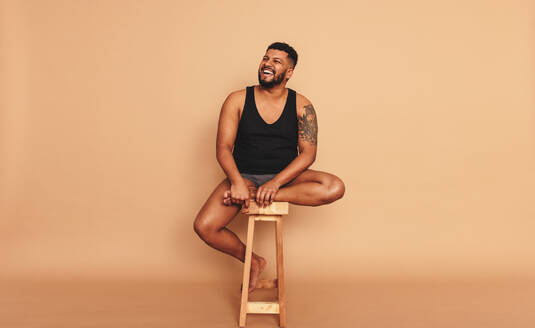 Tattooed man smiling cheerfully while sitting on a wooden chair in a studio. Body positive young man looking away while sitting alone. Self-confident young man feeling comfortable in his natural body. - JLPSF12289