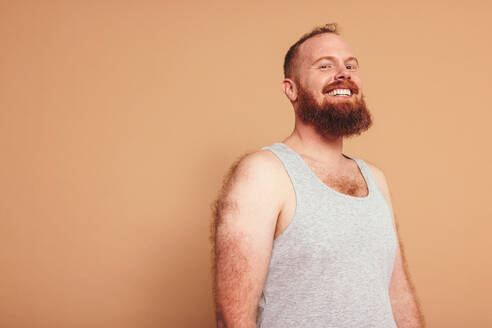 Man with a ginger beard smiling at the camera cheerfully. Body positive young man standing against a studio background. Self-assured young man feeling comfortable in his natural body. - JLPSF12292