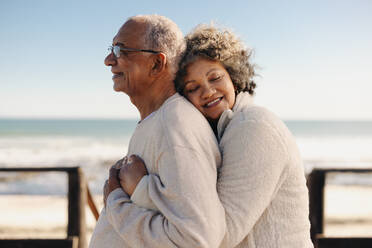 Affectionate senior woman smiling happily while embracing her husband by the ocean. Romantic elderly couple enjoying spending some quality time together after retirement. - JLPSF12304