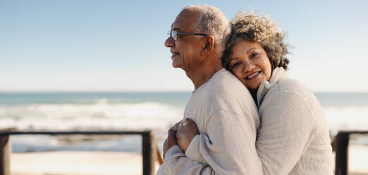 Romantic senior woman smiling at the camera while embracing her husband by the ocean. Affectionate elderly couple enjoying spending some quality time together after retirement. - JLPSF12305