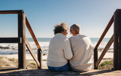 Happy senior couple smiling at each other while sitting on a wooden foot bridge in front of the ocean. Retired elderly couple spending some quality time together at the beach. - JLPSF12308