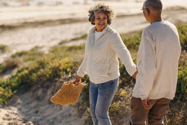 Glückliche ältere Frau, die ihren Mann an der Hand hält, während sie zum Strand geht, um ein Picknick zu machen. Fröhliches älteres Paar, das nach der Pensionierung einen Urlaub am Meer macht. - JLPSF12309