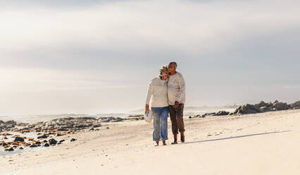 Romantic elderly couple smiling and embracing each other while walking barefoot on beach sand. Happy senior couple enjoying spending some quality time together after retirement. - JLPSF12310