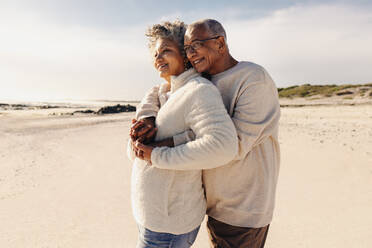 Älteres Paar, das den erfrischenden Blick auf das Meer am Strand genießt. Glückliches älteres Paar, das sich umarmt, während es am Strand steht. Älteres Paar, das seinen Ruhestand zusammen genießt. - JLPSF12314