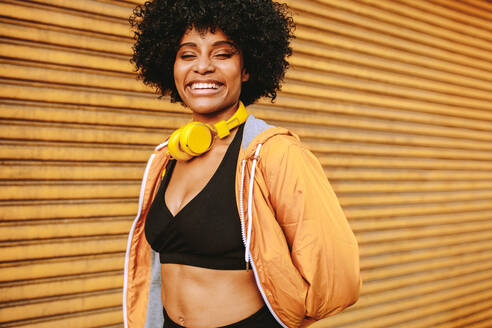 Sporty young woman standing outdoors in the city and smiling. Female athlete with headphones resting after workout. - JLPSF12319