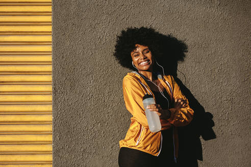 Junge Fitness-Frau steht mit Wasserflasche im Freien und lächelt. Läuferin ruht sich nach dem Training aus und lehnt sich im Freien an eine Wand. - JLPSF12331