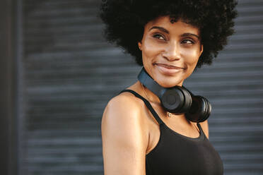 Fit junge Frau mit Kopfhörer lächelnd im Freien. Weibliche Joggerin, die sich nach einem Lauf ausruht, wegschaut und lächelt. - JLPSF12340