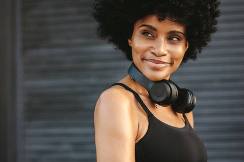 Fit young woman with a headphone smiling outdoors. Female jogger taking rest after a run looking away and smiling. - JLPSF12340