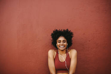 Lächelnde Fitness-Frau, die mit Kopfhörern an einer Wand steht. Frau in Trainingskleidung entspannt sich nach ihrem Training. - JLPSF12355