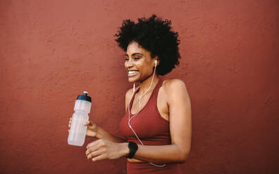 Fitness-Frau mit Wasserflasche stehend und lächelnd. Läuferin ruht sich nach dem Training aus, lehnt sich an eine Wand, schaut weg und lächelt. - JLPSF12361