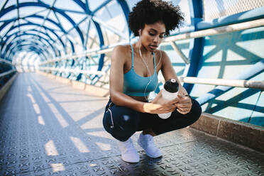 Female in sportswear crouching on a bridge with a water bottle. Young woman runner resting after running exercise in the city. - JLPSF12373