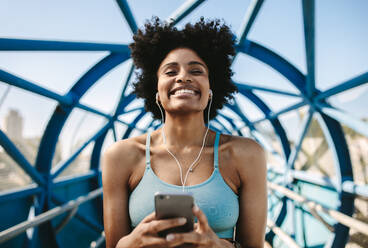 Happy female runner enjoying listening to music from her phone after run. African female relaxing after workout. - JLPSF12381