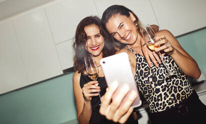 Cheerful women friends partying at home drinking wine. Women taking a selfie using a mobile phone holding wine glasses. - JLPSF12387