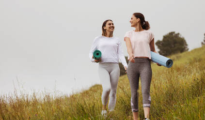Lächelnde Freundinnen in Fitnesskleidung, die mit Yogamatten über die Wiesen spazieren. Zwei Fitnessfrauen beim Workout im Freien. - JLPSF12399